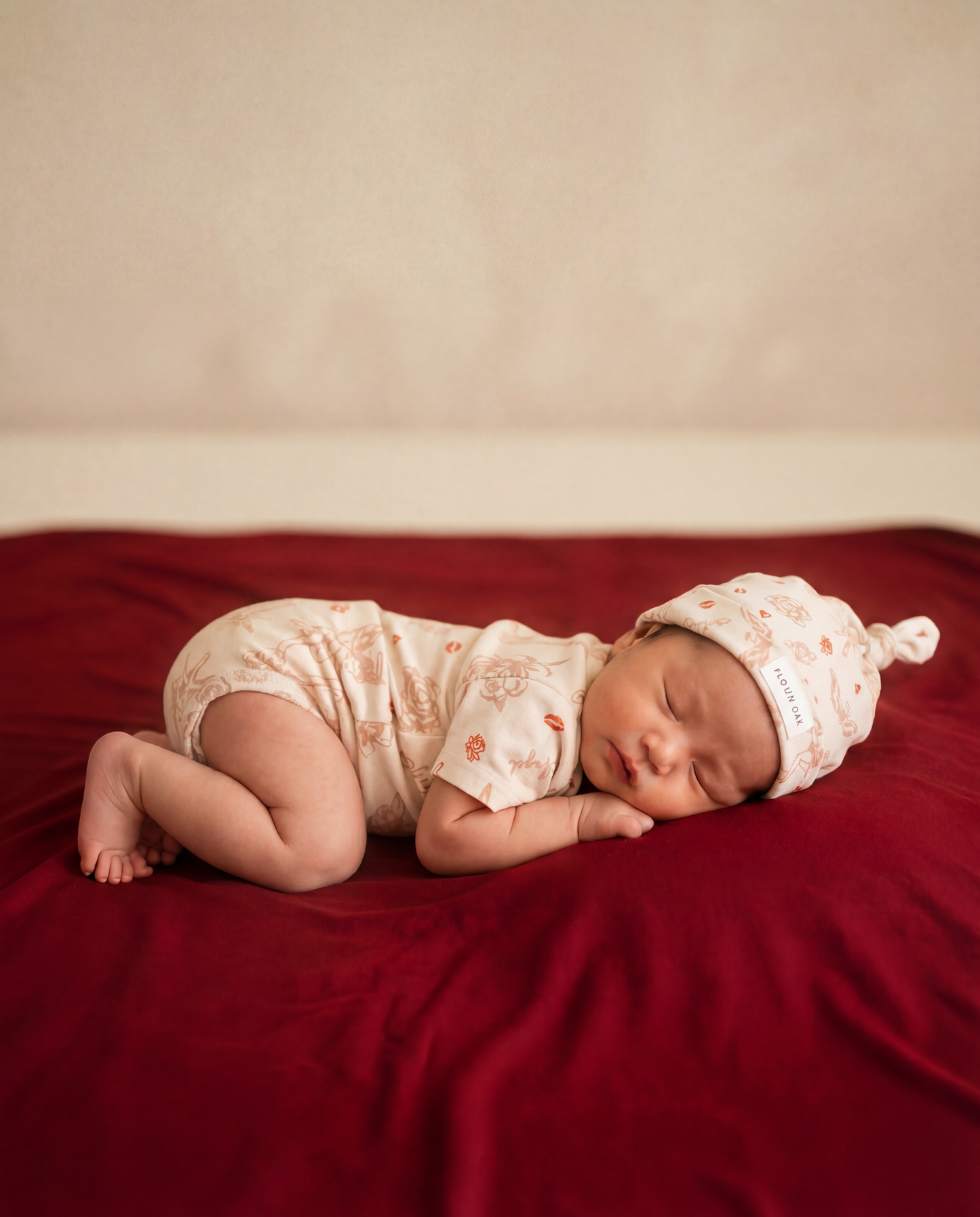 Newborn baby sleeping on a red blanket wearing a white outfit with floral patterns and a matching hat.