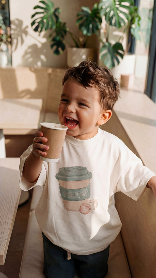 Child wearing a white t-shirt with a coffee cup design, holding a paper cup.