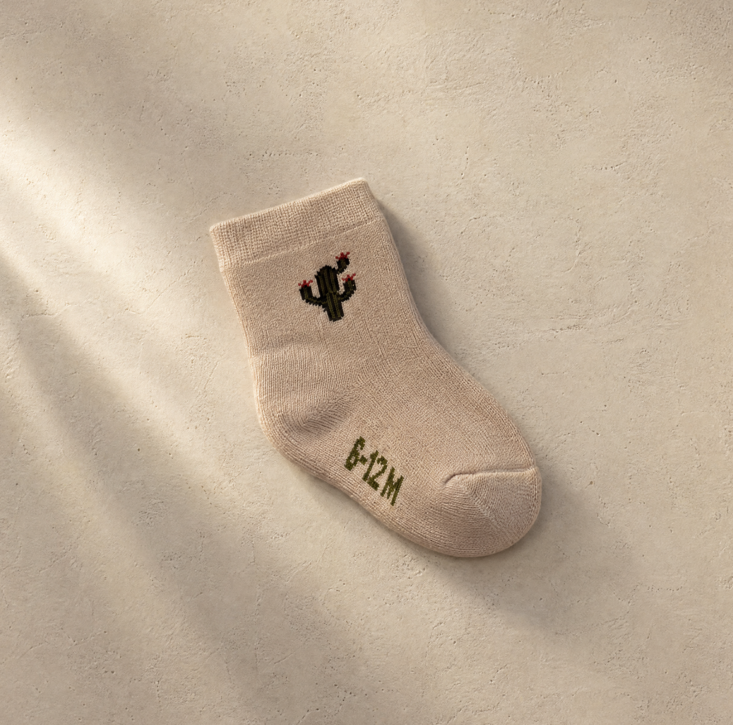 A single beige baby sock with a small embroidered black cactus and the text '6+12M' in olive green, displayed on a textured beige background with soft diagonal shadows.
