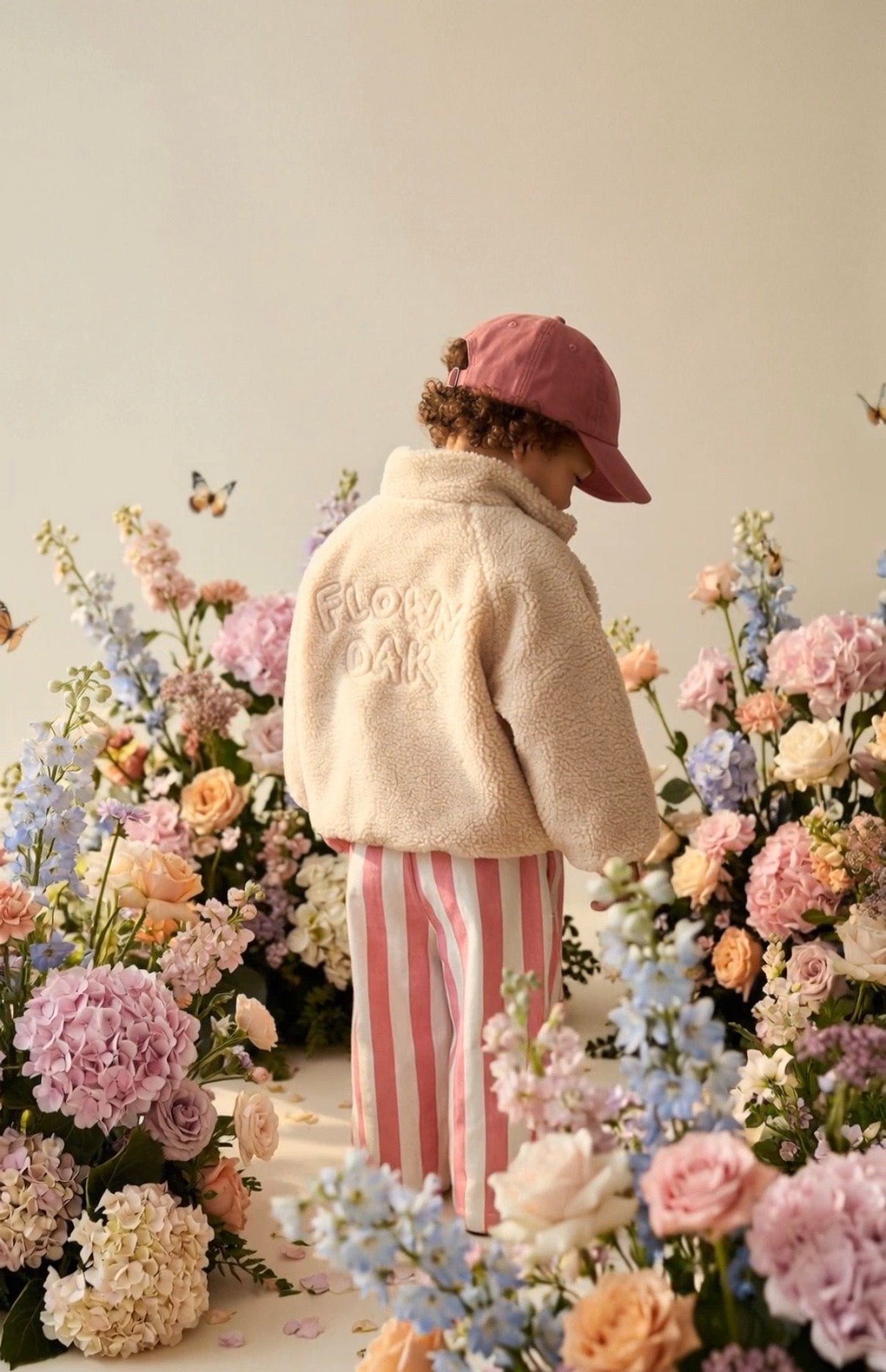 A child wearing a beige puffer jumper and pink cap among colorful flowers indoors.