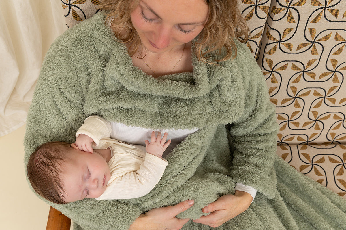 Woman holding a baby wrapped in a green wrap against a patterned wall.