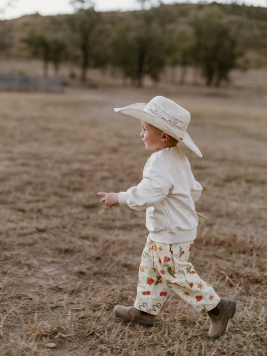 A toddler walking outdoors in a dry field, wearing a cream cowboy hat, cream sweatshirt, cream pants with a red and green floral pattern, and brown boots.