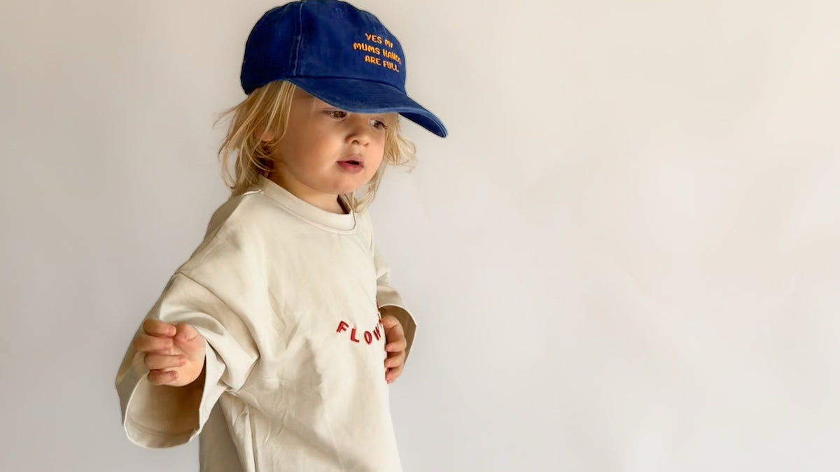 A toddler standing against a plain light background, wearing a cream-colored oversized t-shirt with red embroidered text, blue jeans, and a blue baseball cap.