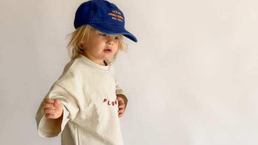 A toddler standing against a plain light background, wearing a cream-colored oversized t-shirt with red embroidered text, blue jeans, and a blue baseball cap.