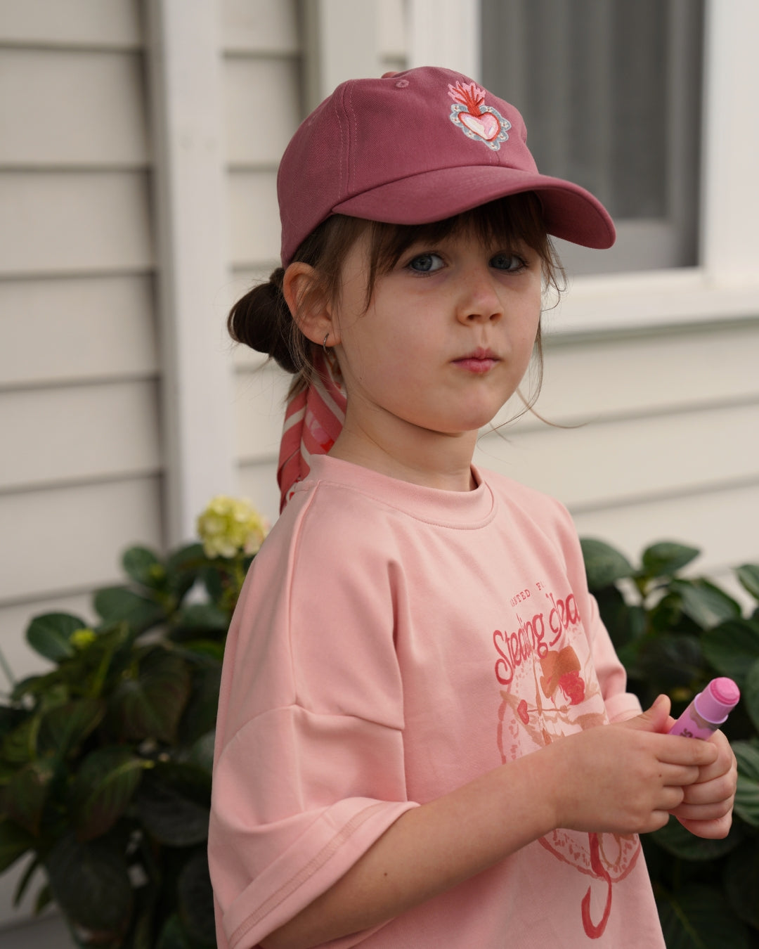 Young girl wearing a pink cap and shirt, holding a pink object outdoors.