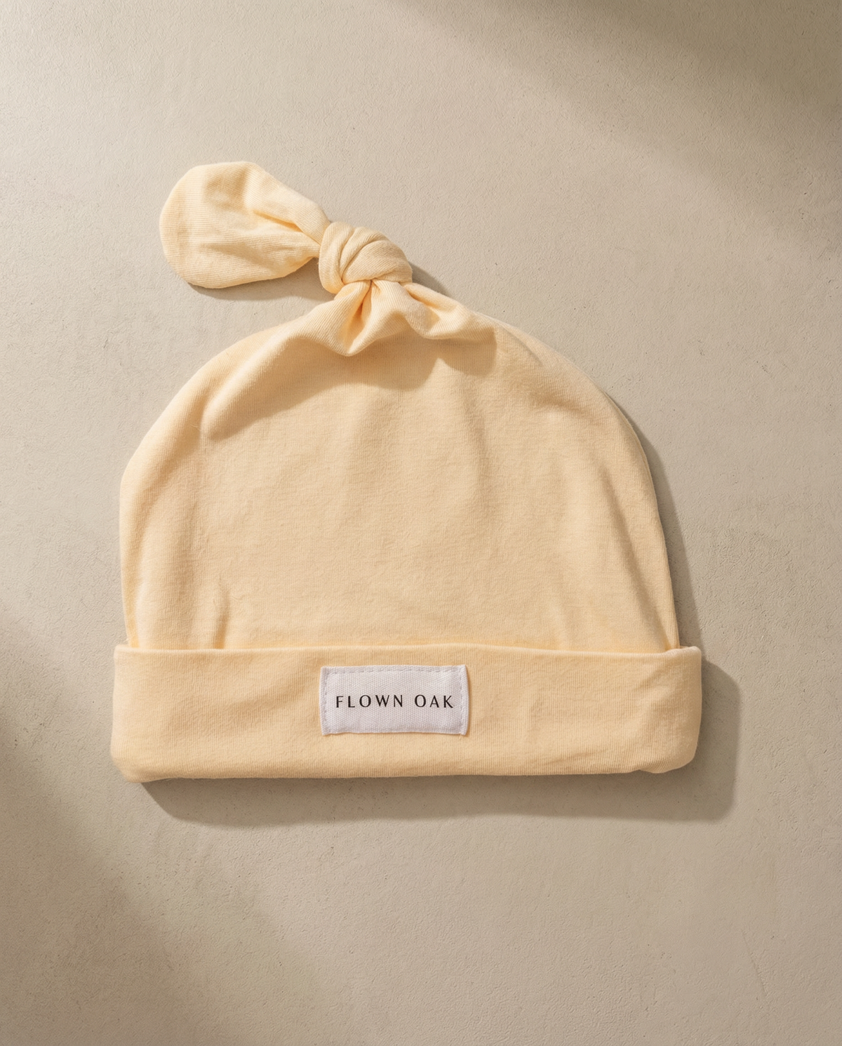 Beige baby hat with a knot on top and 'Flown Oak' label on a beige background