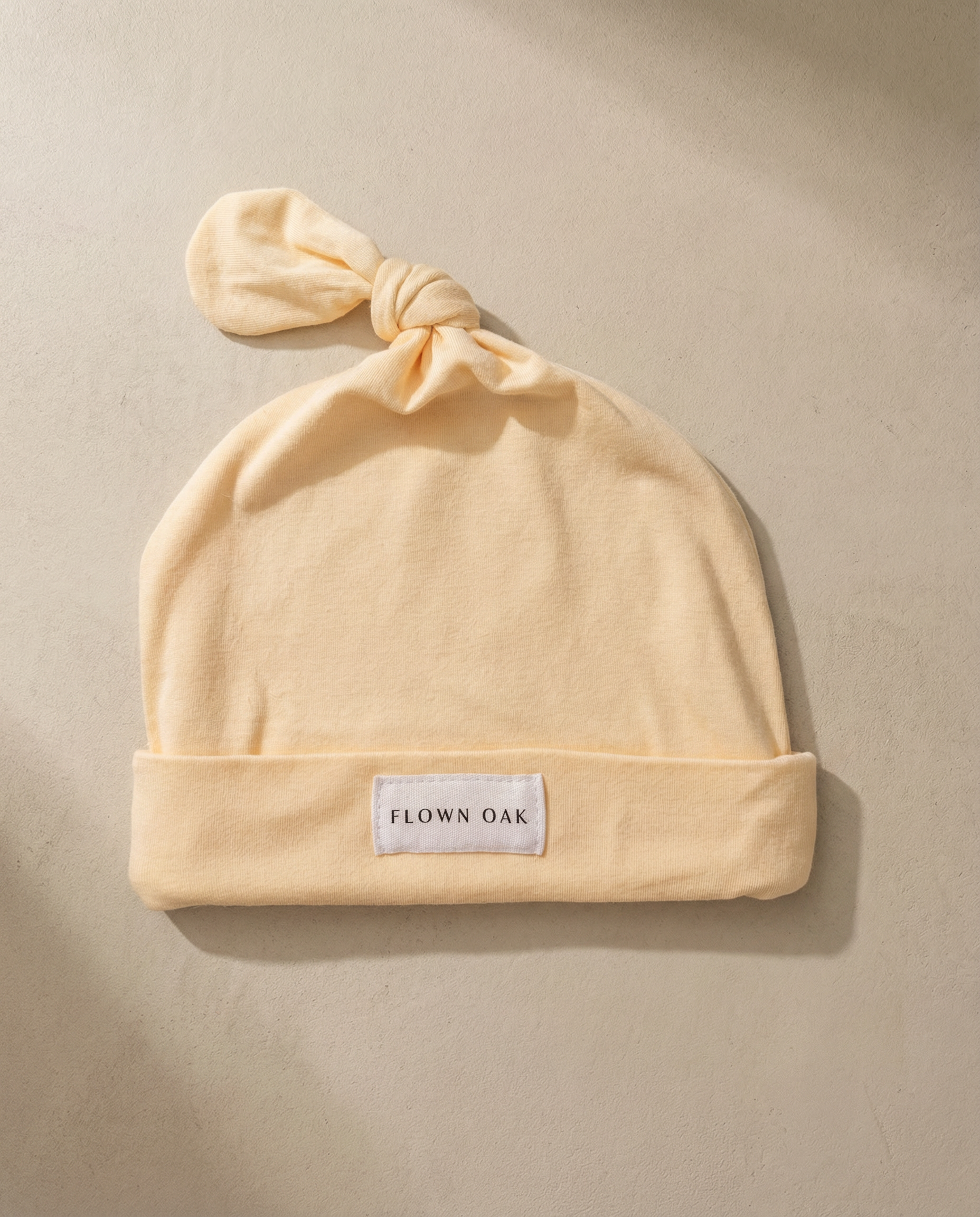 Beige baby hat with a knot on top and 'Flown Oak' label on a beige background