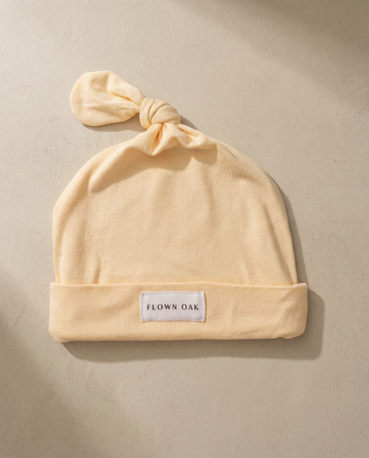 Beige baby hat with a knot on top and 'Flown Oak' label on a beige background