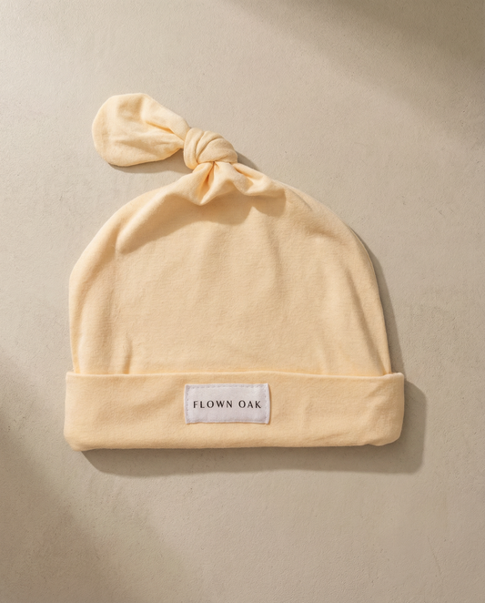 Beige baby hat with a knot on top and 'Flown Oak' label on a beige background