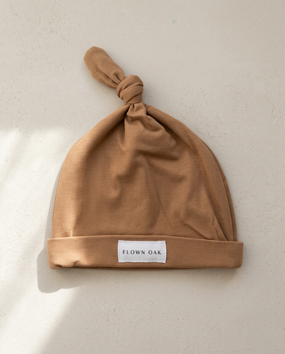 Brown baby hat with a knotted top on a light beige background, featuring the brand 'Flown Oak'.