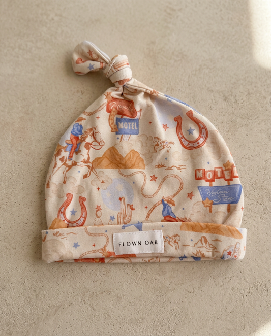 Baby hat with cowboy design and 'Flown Oak' label on a beige background