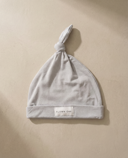 Gray baby hat with a knot on top, featuring a 'Flown Oak' label, on a beige background.