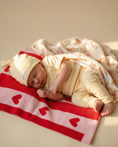 Newborn baby sleeping on a red blanket with heart patterns, wrapped in a white blanket.
