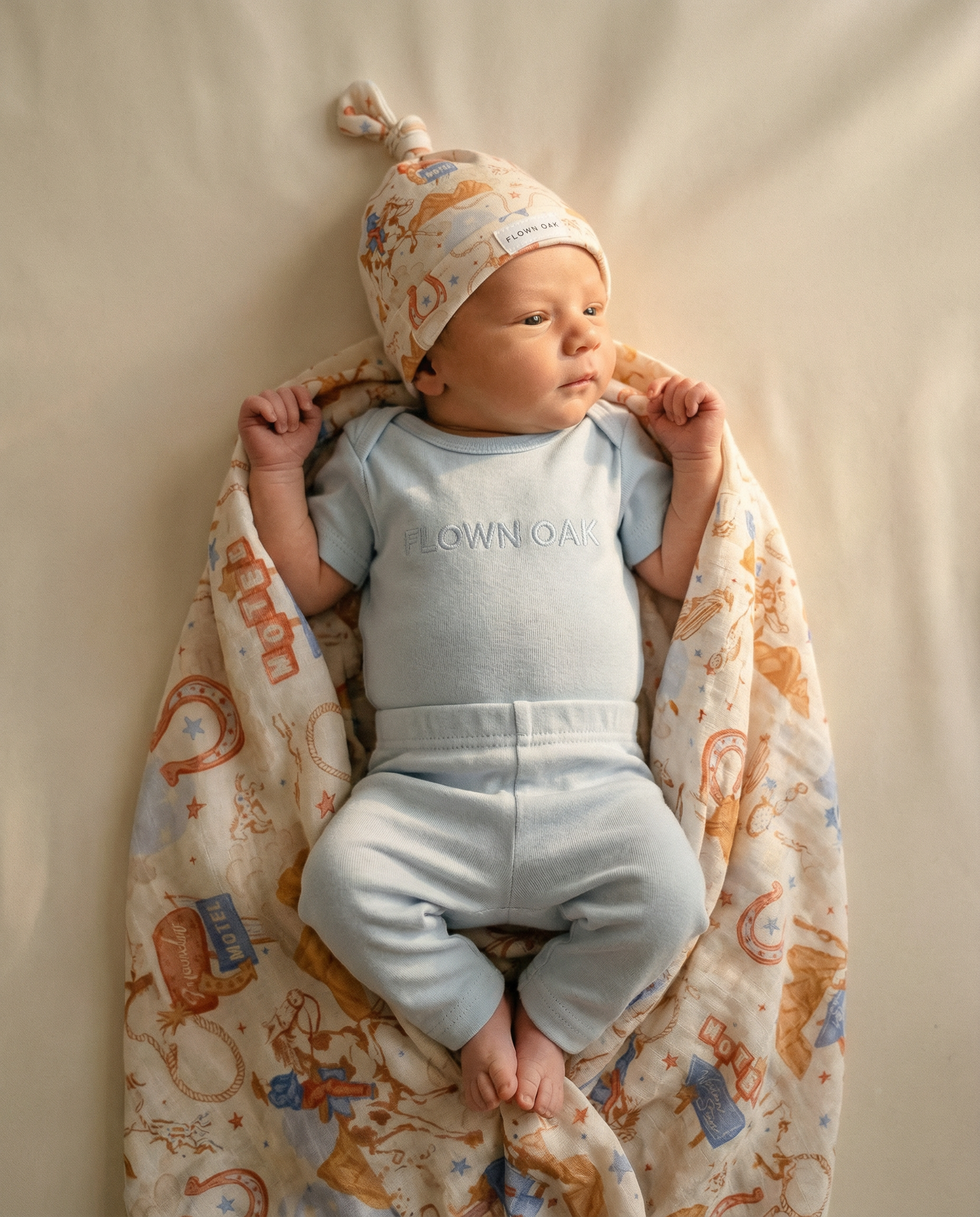 Baby in light blue outfit with 'DOWNNOWN' text, wrapped in a patterned blanket on a beige background