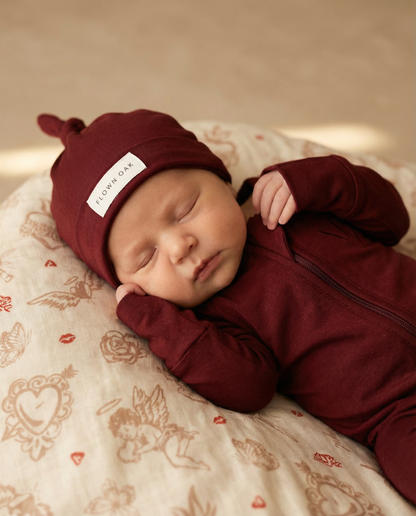 Newborn baby in a maroon outfit and hat sleeping on a patterned blanket.