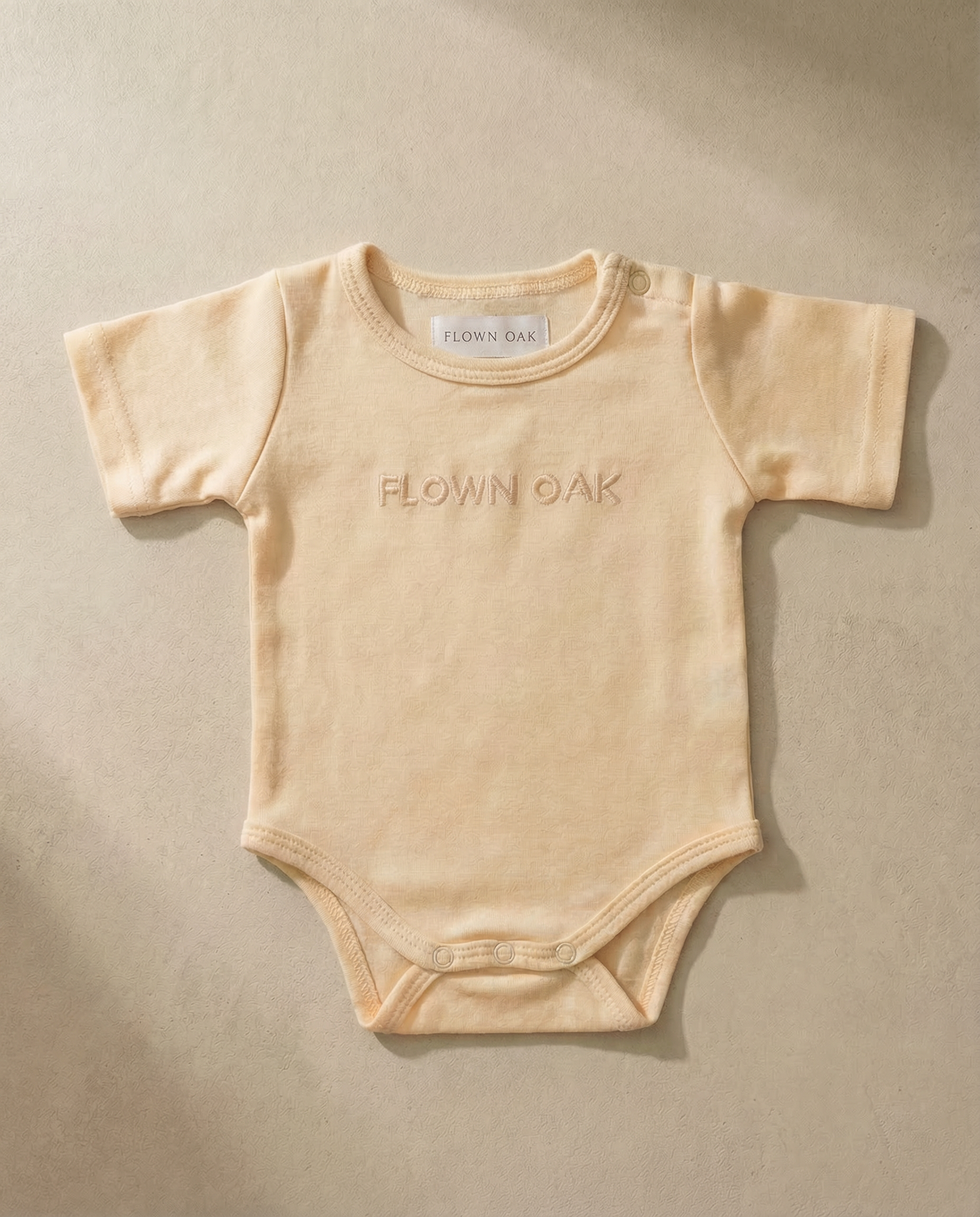 Beige baby onesie with 'Flown Oak' branding on a beige background