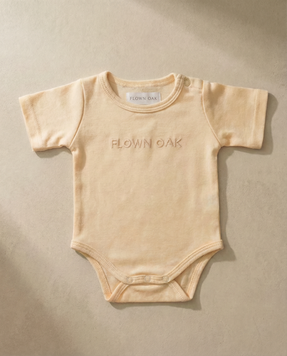 Beige baby onesie with 'Flown Oak' branding on a beige background
