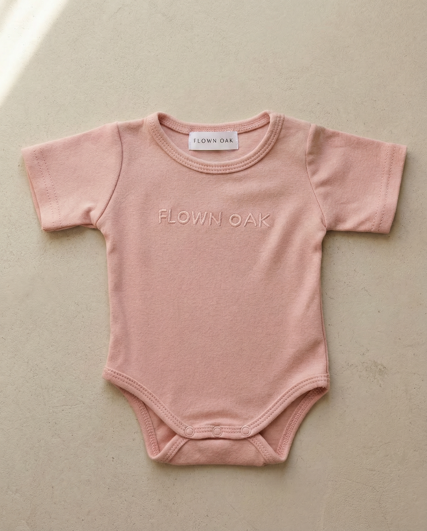 Pink baby onesie with 'Flown Oak' branding on a beige background