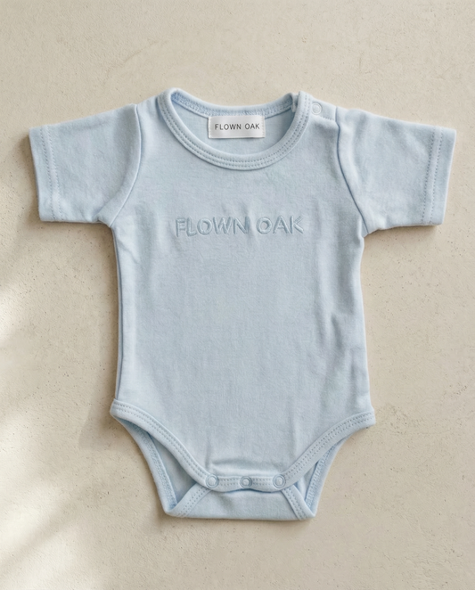 Light blue baby onesie with 'Flown Oak' branding on a beige background