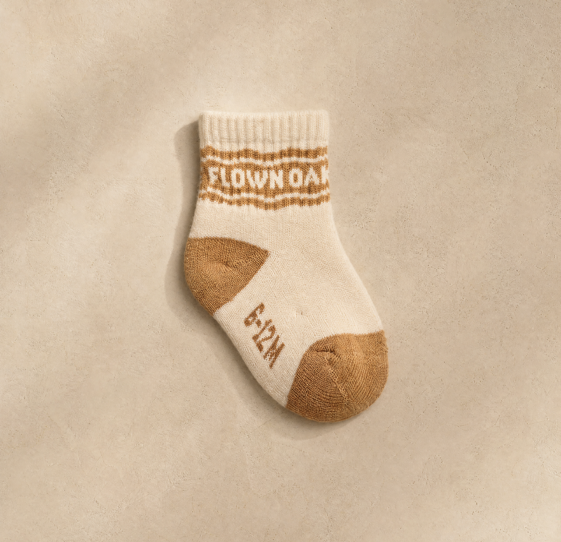 A single baby sock in cream and tan, laid flat on a textured beige background. The sock features the words 'FLOWER OAK' in tan across the ribbed cuff and '6-12M' on the side.
