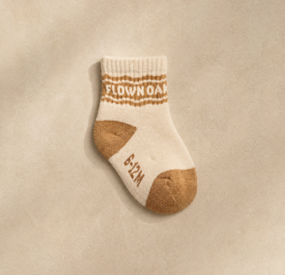 A single baby sock in cream and tan, laid flat on a textured beige background. The sock features the words 'FLOWER OAK' in tan across the ribbed cuff and '6-12M' on the side.