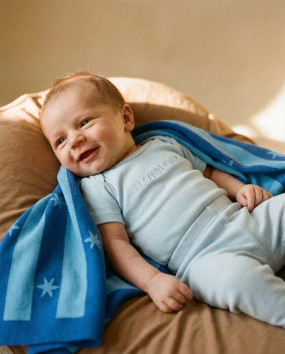 Baby lying on a bed with a blue blanket, wearing a light blue onesie.