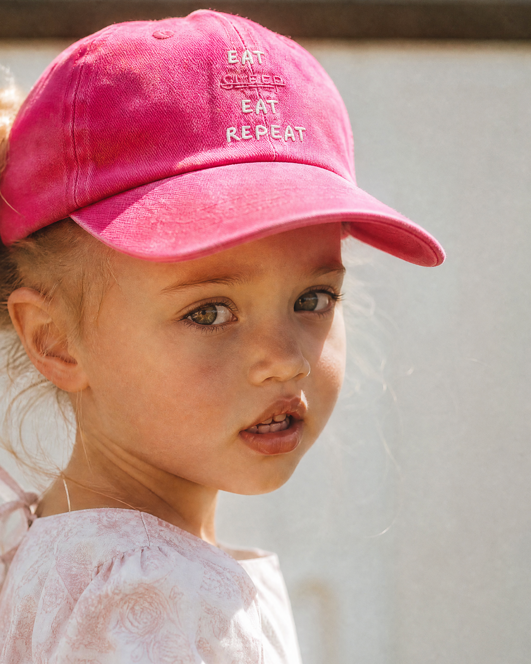 Child wearing a pink cap with text, standing against a light-colored wall.