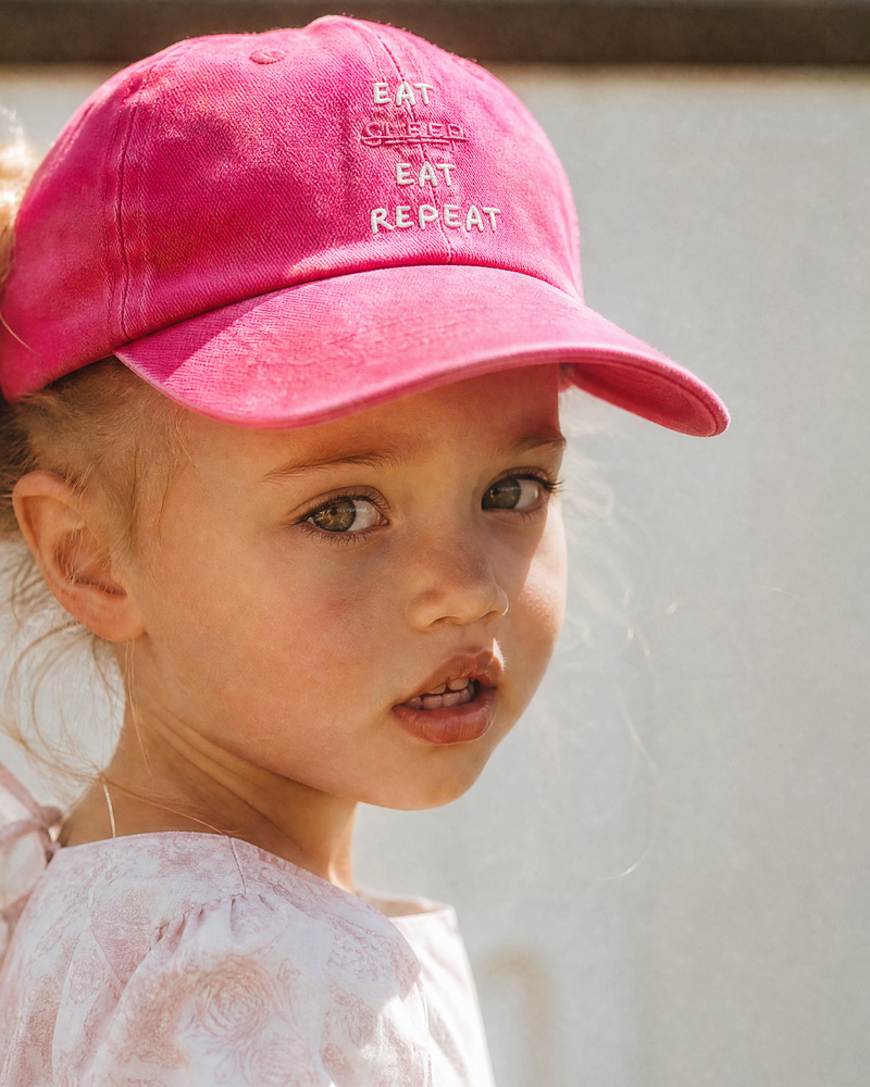 Child wearing a pink cap with text, standing against a light-colored wall.