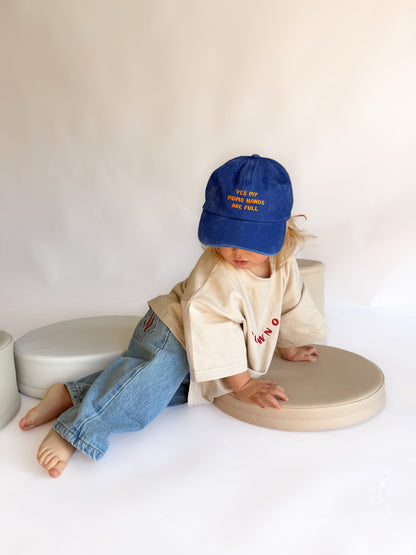 A toddler sits on round beige cushions in a studio setting, wearing a blue baseball cap with yellow embroidered text, a cream t-shirt, and blue jeans.