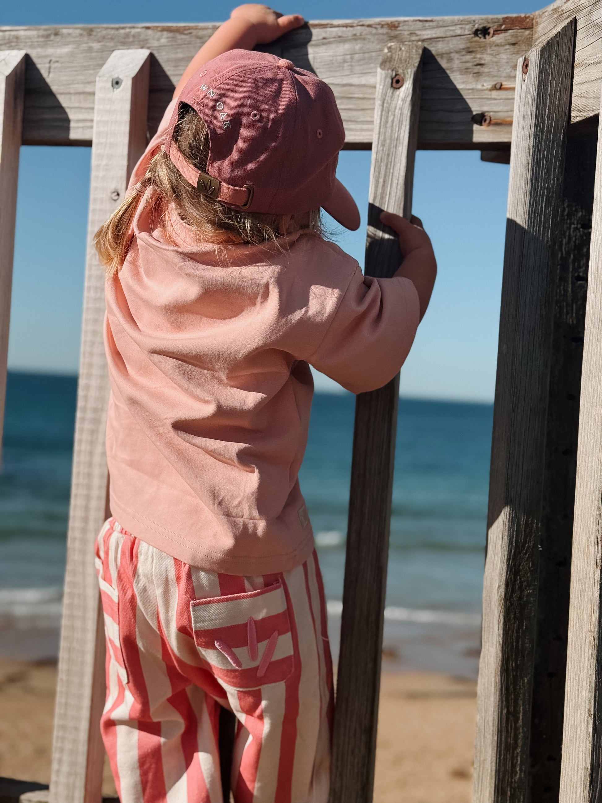 Child wearing a pink cap and outfit standing on a wooden structure by the beach.