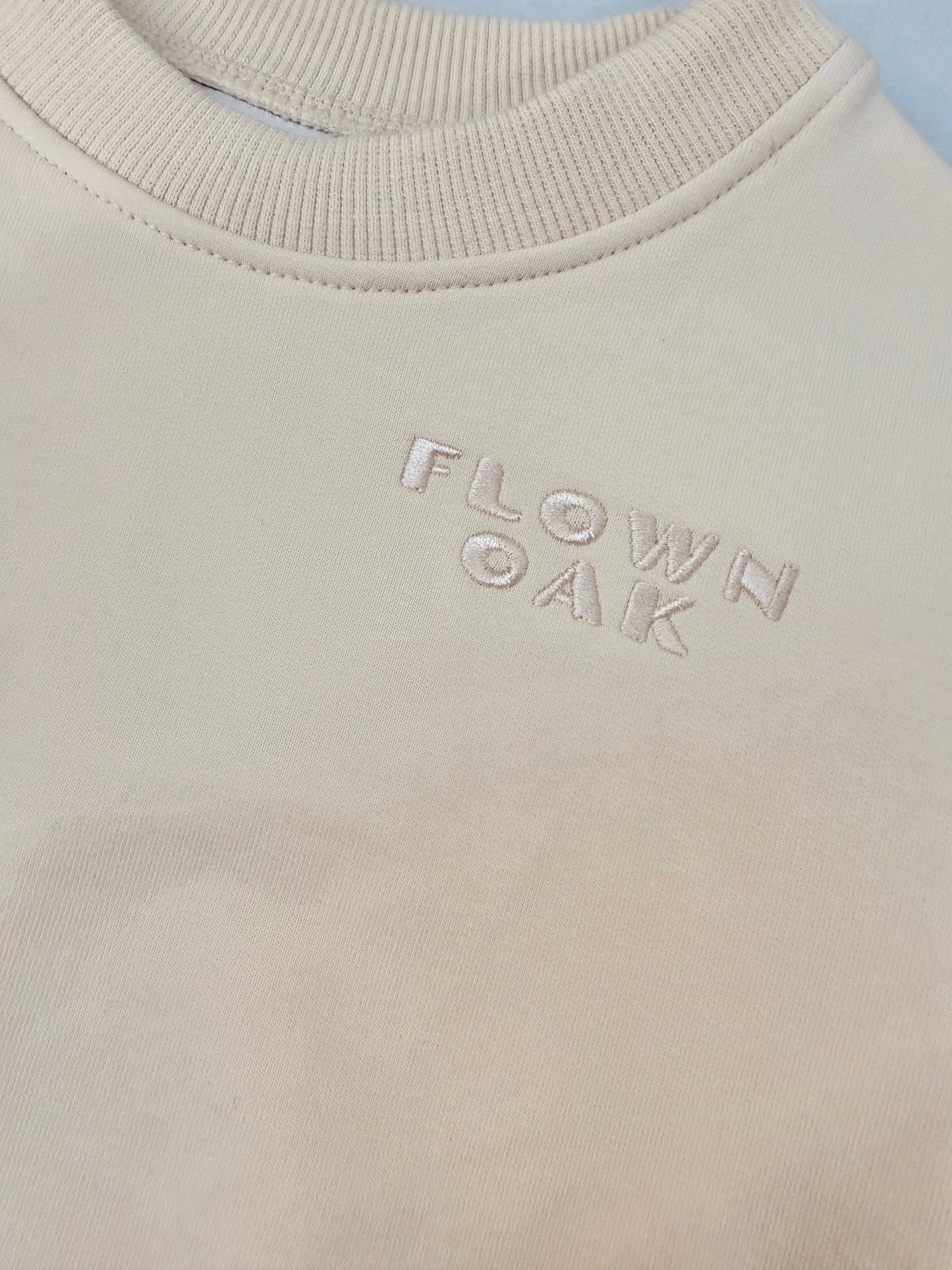 Close-up of a cream-colored crewneck sweatshirt with the brand name 'FLOWN OAK' subtly embroidered on the chest.