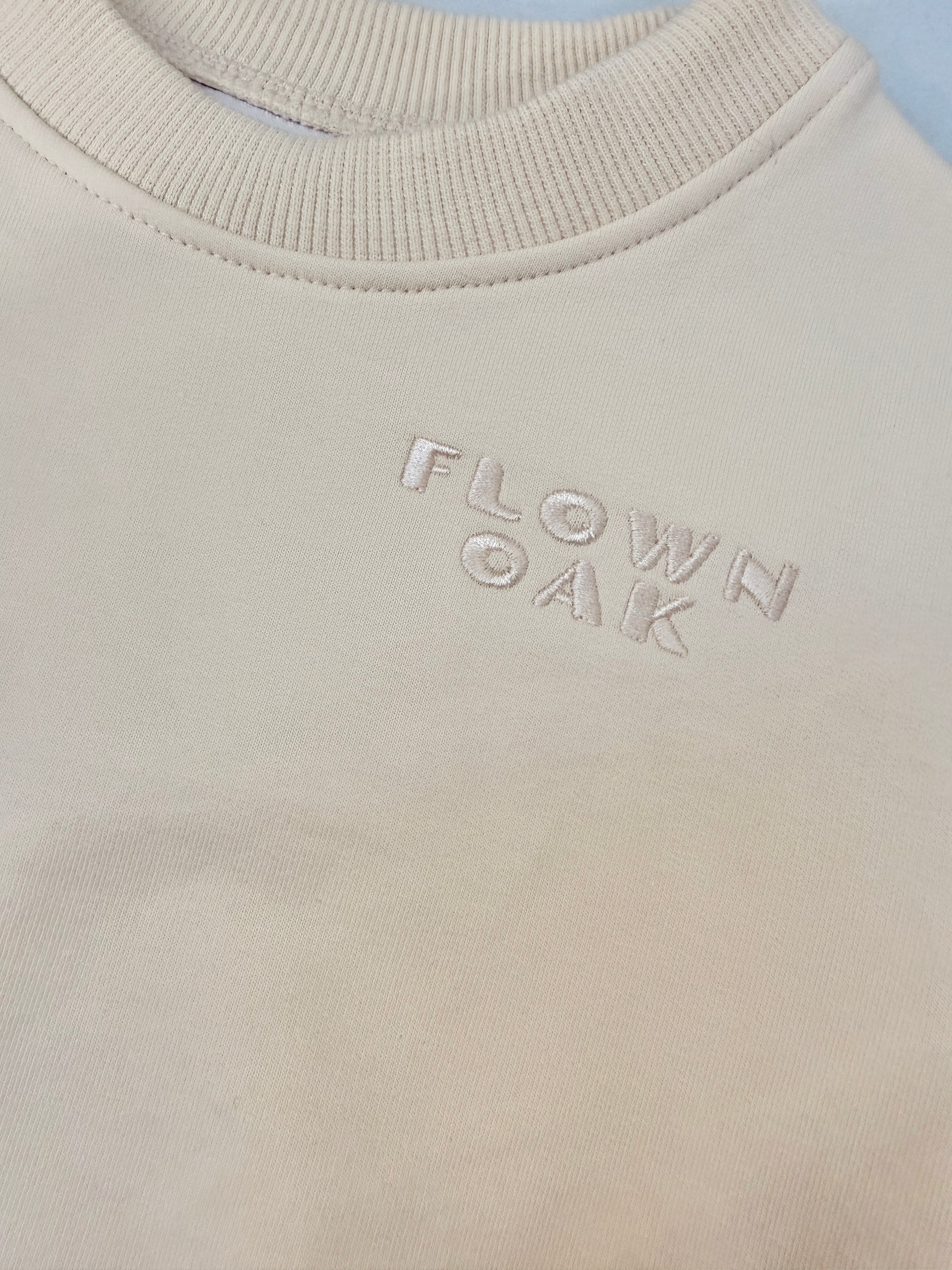 Close-up of a cream-colored crewneck sweatshirt with the brand name 'FLOWN OAK' subtly embroidered on the chest.