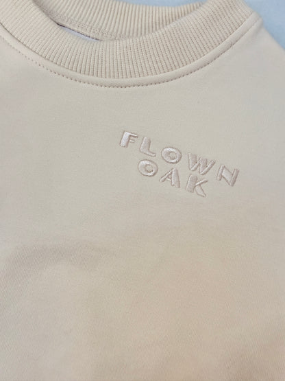 Close-up of a cream-colored crewneck sweatshirt with the brand name 'FLOWN OAK' subtly embroidered on the chest.