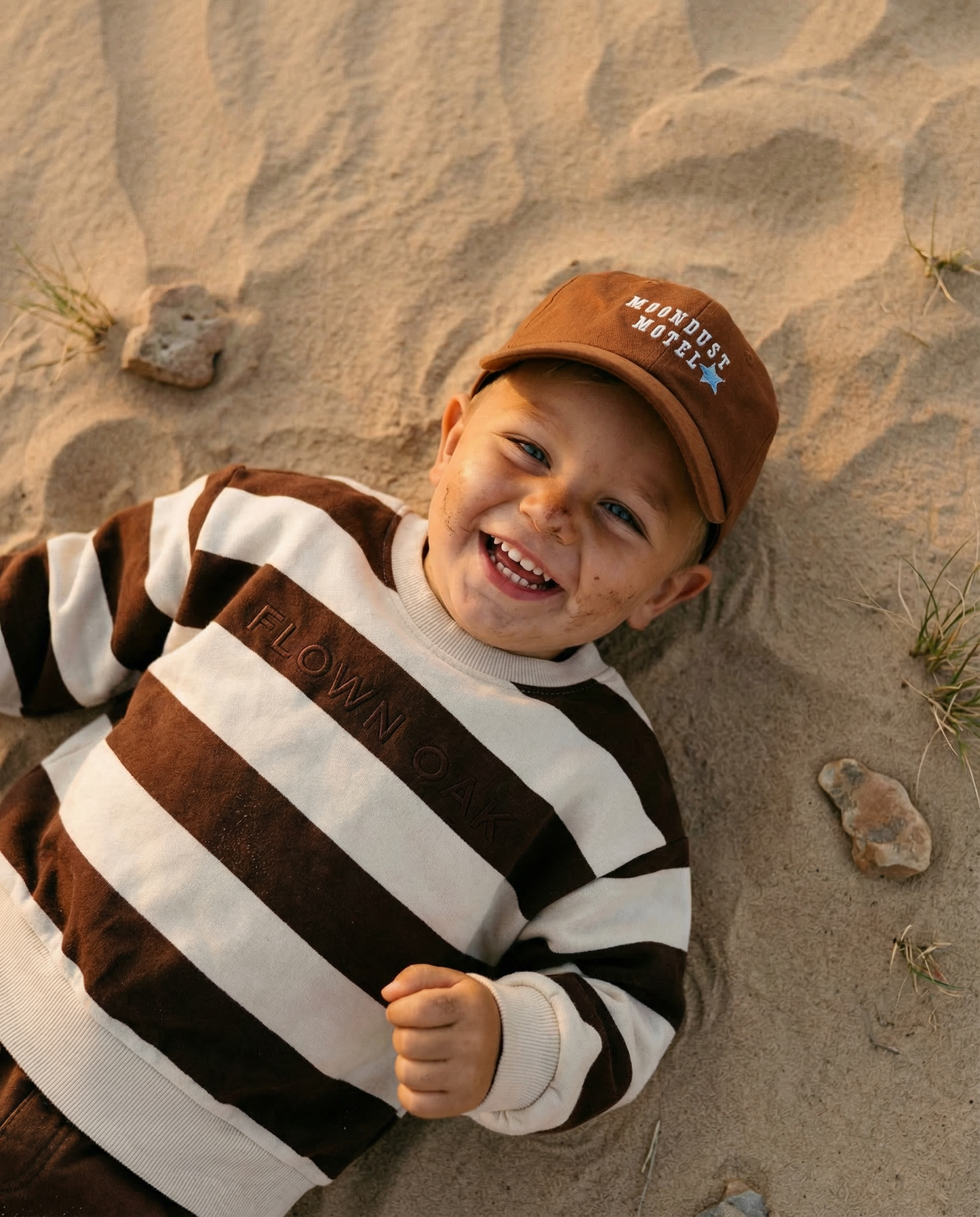 Child wearing a brown and white striped sweater and cap, lying on sand with a smile.