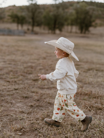 A toddler walking outdoors in a dry field, wearing a cream cowboy hat, cream sweatshirt, cream pants with a red and green floral pattern, and brown boots.
