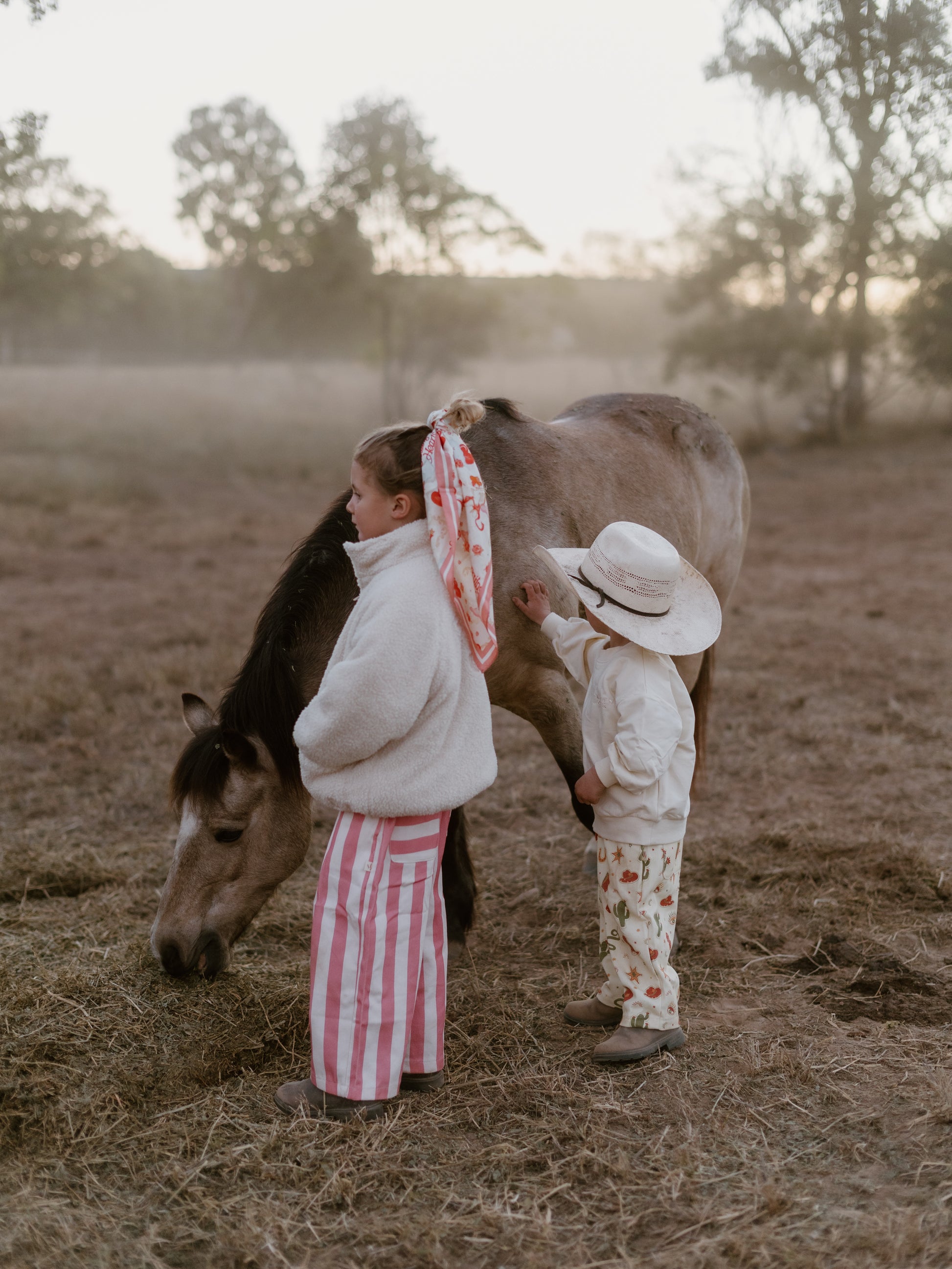 Two children in matching cream and pink patterned outfits stand in a dry field next to a horse at sunset. One child wears a white jacket and pink-and-white striped pants, and the other wears a cream outfit with a strawberry print and a white sun hat.