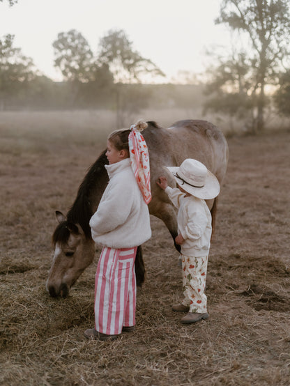 Two children in matching cream and pink patterned outfits stand in a dry field next to a horse at sunset. One child wears a white jacket and pink-and-white striped pants, and the other wears a cream outfit with a strawberry print and a white sun hat.
