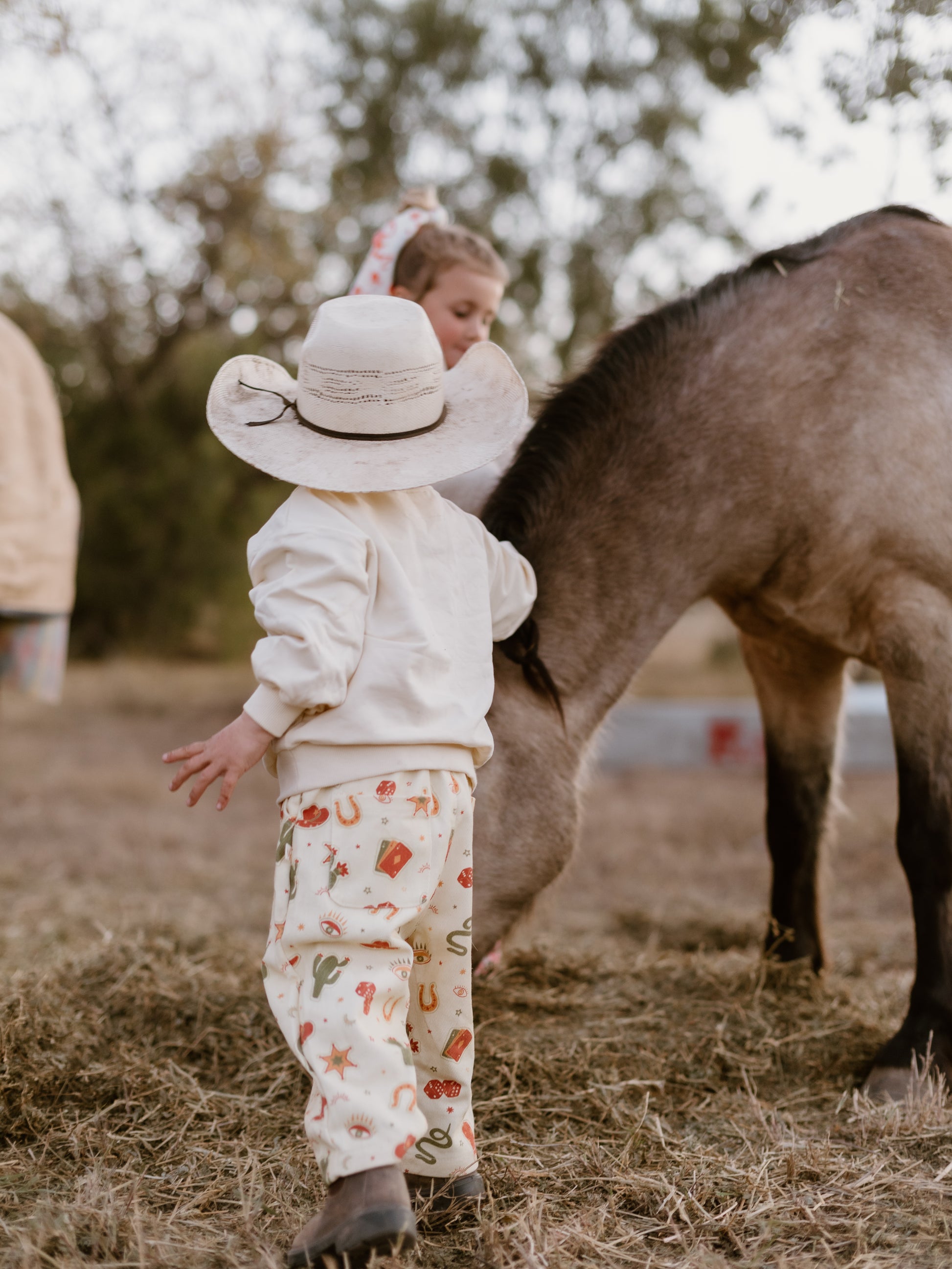 A toddler stands in a field with a horse, wearing a cream wide-brim hat, cream sweatshirt, and cream pants with a small red and green pattern. A woman is standing behind the child.