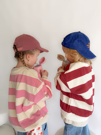 Two young children standing side by side against a white wall, wearing oversized striped sweatshirts and baseball caps. The child on the left wears a pink and cream striped sweatshirt with a dusty rose cap, and the child on the right wears a red and cream striped sweatshirt with a blue cap.
