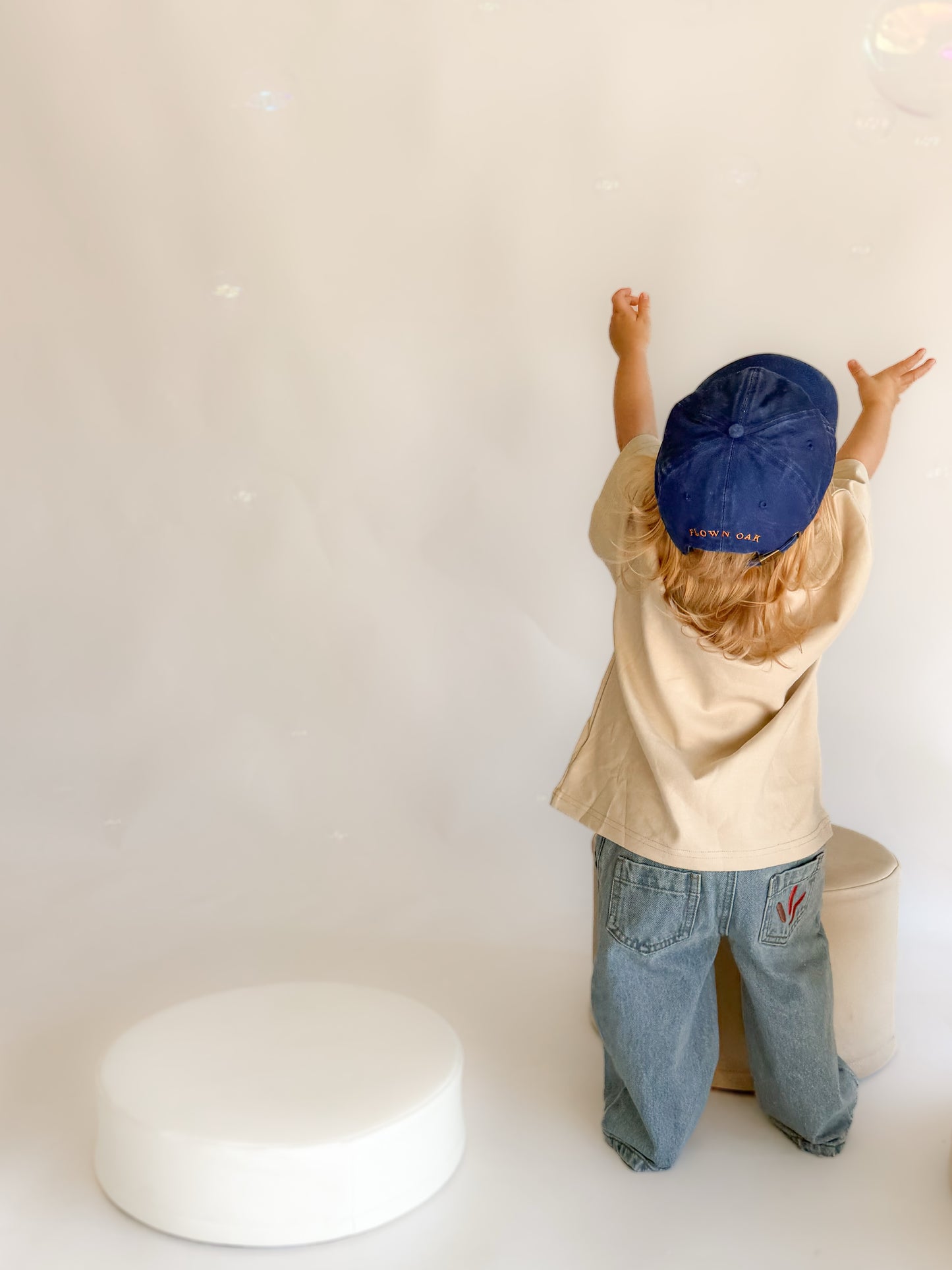 A child seen from behind wearing a beige shirt, blue jeans, and a navy blue baseball cap, standing in a bright studio with white props against a white background.