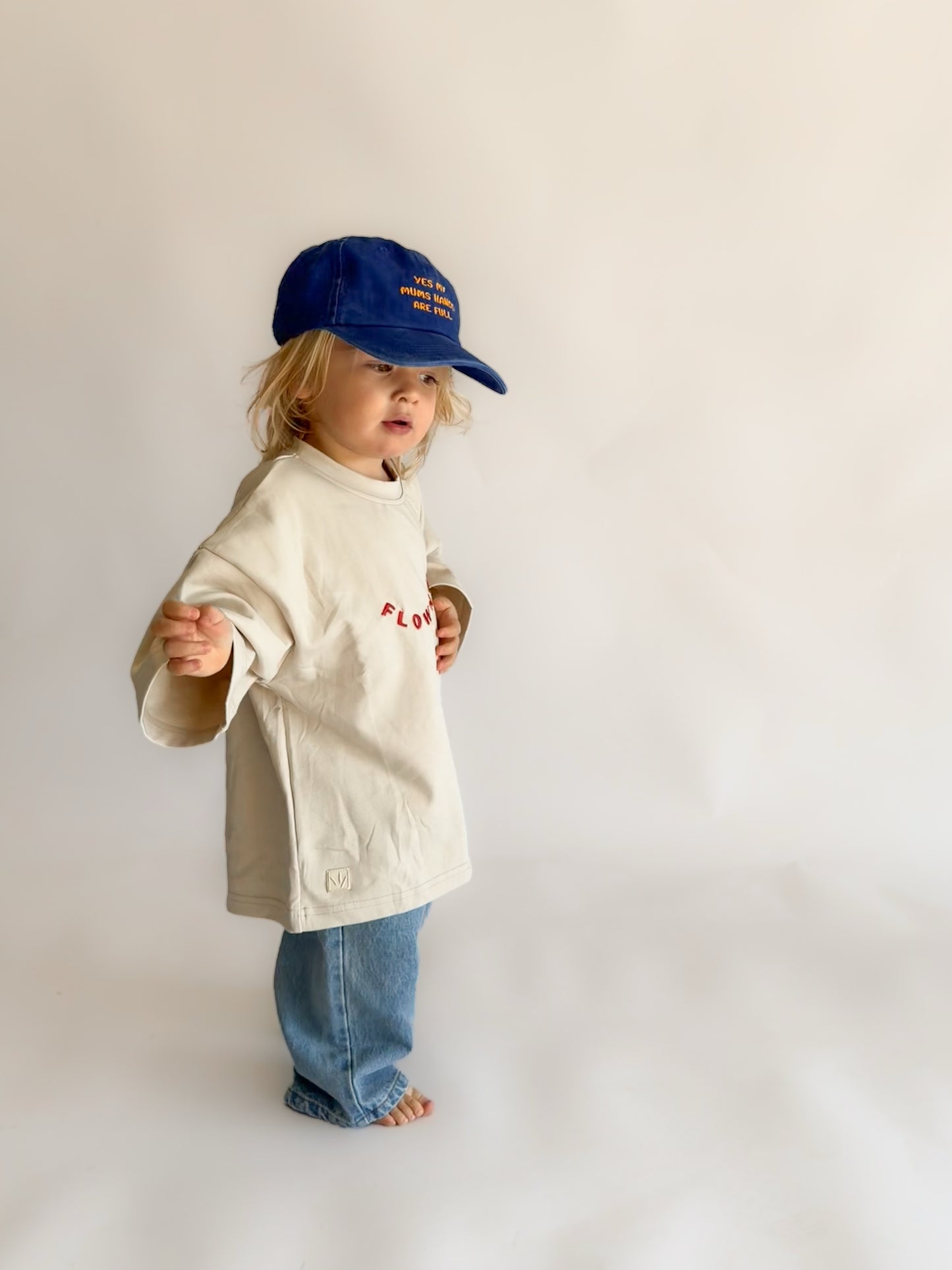 A toddler standing against a plain light background, wearing a cream-colored oversized t-shirt with red embroidered text, blue jeans, and a blue baseball cap.