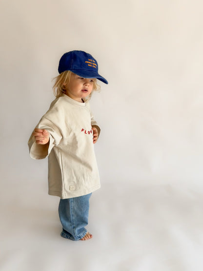 A toddler standing against a plain light background, wearing a cream-colored oversized t-shirt with red embroidered text, blue jeans, and a blue baseball cap.