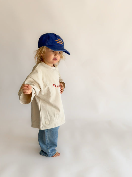 A toddler standing against a plain light background, wearing a cream-colored oversized t-shirt with red embroidered text, blue jeans, and a blue baseball cap.