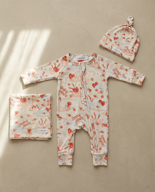 Baby outfit set with romper, hat, and blanket featuring heart and bunny designs on a beige background.