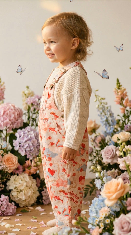 Child wearing heart overalls standing among flowers with butterflies.