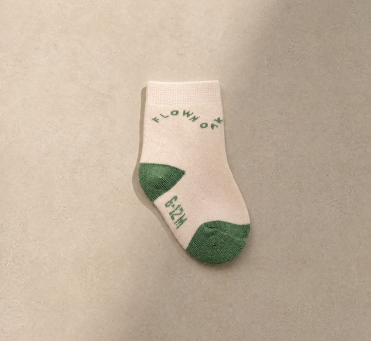 A single off-white knit sock with green heel and toe accents, featuring the words 'FLOW OF LIFE' in green on the side, displayed on a neutral beige textured background.